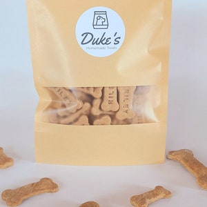 May include: A brown paper bag with a white label that says "Duke's Homemade Treats". The bag is filled with dog treats shaped like bones. The treats are labeled "Riley".