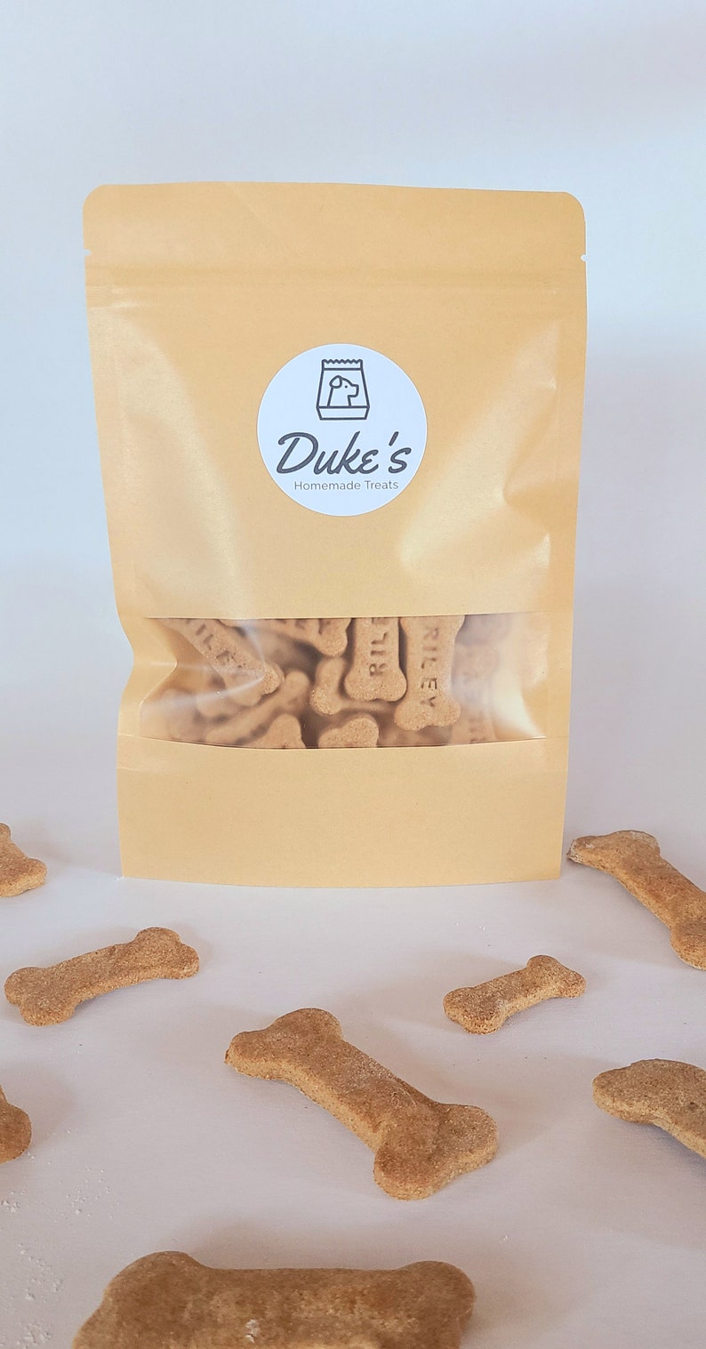 May include: A brown paper bag with a white label that says "Duke's Homemade Treats". The bag is filled with dog treats shaped like bones. The treats are labeled "Riley".