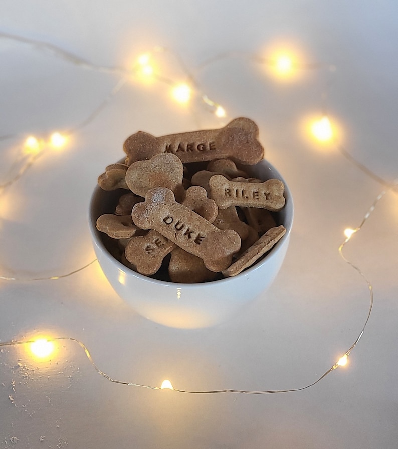 May include: A white bowl filled with homemade dog treats shaped like bones. The treats are labeled with the names "Duke", "Marge", and "Riley".