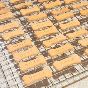 May include: A close-up of a wire rack with multiple dog bone-shaped treats. The treats are light brown and have the word "Duke" written on them.