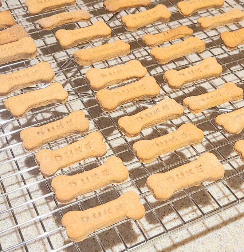 May include: A close-up of a wire rack with multiple dog bone-shaped treats. The treats are light brown and have the word "Duke" written on them.