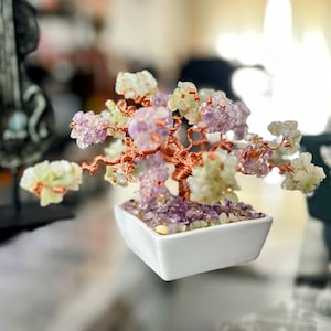 May include: A decorative bonsai tree with a copper wire trunk and branches, featuring amethyst and other colourful gemstone leaves. The tree sits in a square white ceramic pot filled with amethyst chips, a unique home decor item.