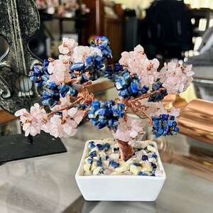 Op de afbeelding: Een roze en blauwe kristalboom van draad in een witte keramische pot. De boom is gemaakt van rozenkwarts- en lapis lazuli-kristallen.