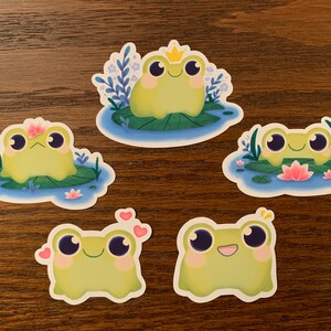 May include: Five frog stickers with different expressions. Each frog is sitting on a lily pad with flowers and leaves. The frogs are green with big eyes and smiles. One frog is wearing a crown.