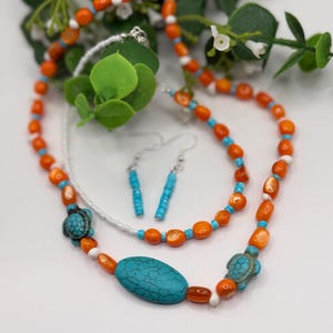 Puede incluir: Un conjunto de joyería compuesto por un collar y pendientes a juego. El collar presenta cuentas naranjas y blancas, dos dijes de tortuga de color turquesa y una cuenta grande ovalada de color turquesa. Los pendientes están hechos de pequeñas cuentas turquesas.