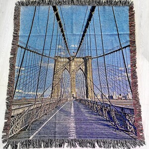 100% woven throw blanket with a photographic image of the Brooklyn Bridge in NYC. The image features a perspective view from the center of the footpath with an American flag on top of the bridge. The blanket has a colorful fringe on all sides.