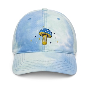 May include: A blue and green tie-dye baseball cap with an embroidered blue and yellow mushroom design. The mushroom has white spots and is surrounded by small yellow stars.