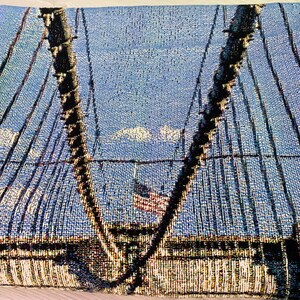 100% woven throw blanket with a photographic image of the Brooklyn Bridge in NYC. The image features a perspective view from the center of the footpath with an American flag on top of the bridge. The blanket has a colorful fringe on all sides.