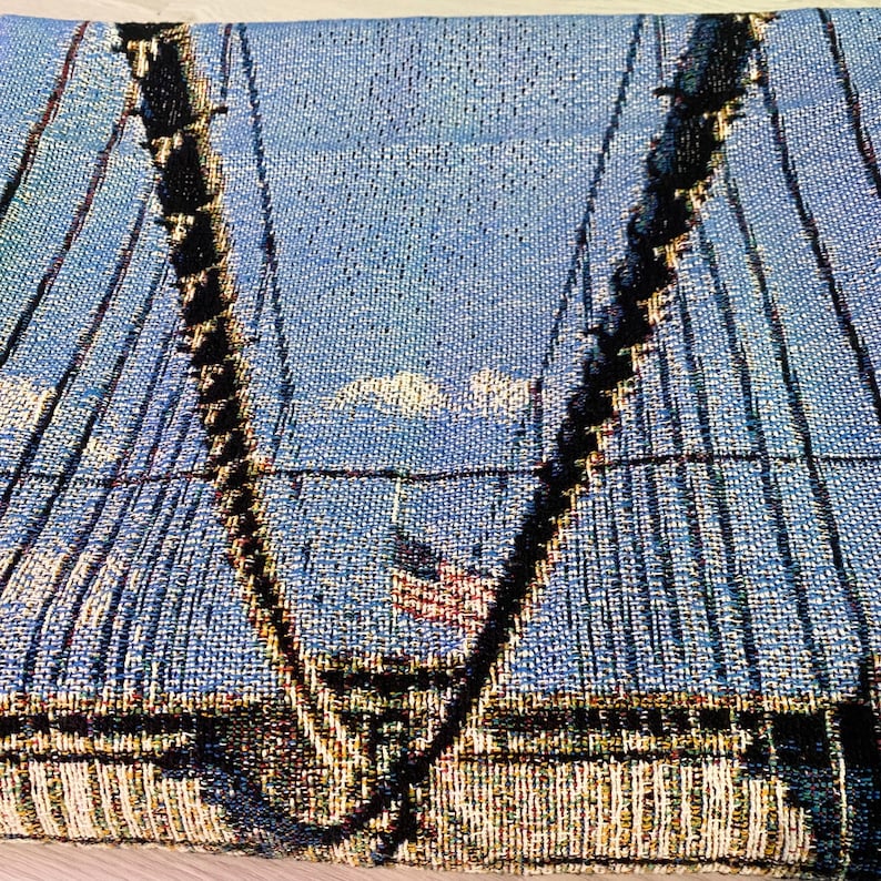100% woven throw blanket with a photographic image of the Brooklyn Bridge in NYC. The image features a perspective view from the center of the footpath with an American flag on top of the bridge. The blanket has a colorful fringe on all sides.