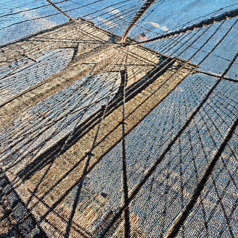 100% woven throw blanket with a photographic image of the Brooklyn Bridge in NYC. The image features a perspective view from the center of the footpath with an American flag on top of the bridge. The blanket has a colorful fringe on all sides.