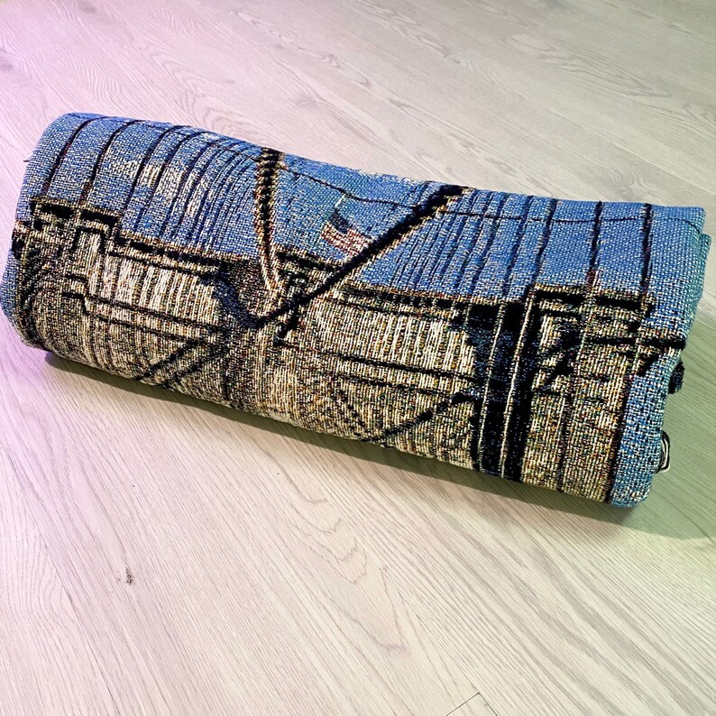 100% woven throw blanket with a photographic image of the Brooklyn Bridge in NYC. The image features a perspective view from the center of the footpath with an American flag on top of the bridge. The blanket has a colorful fringe on all sides.