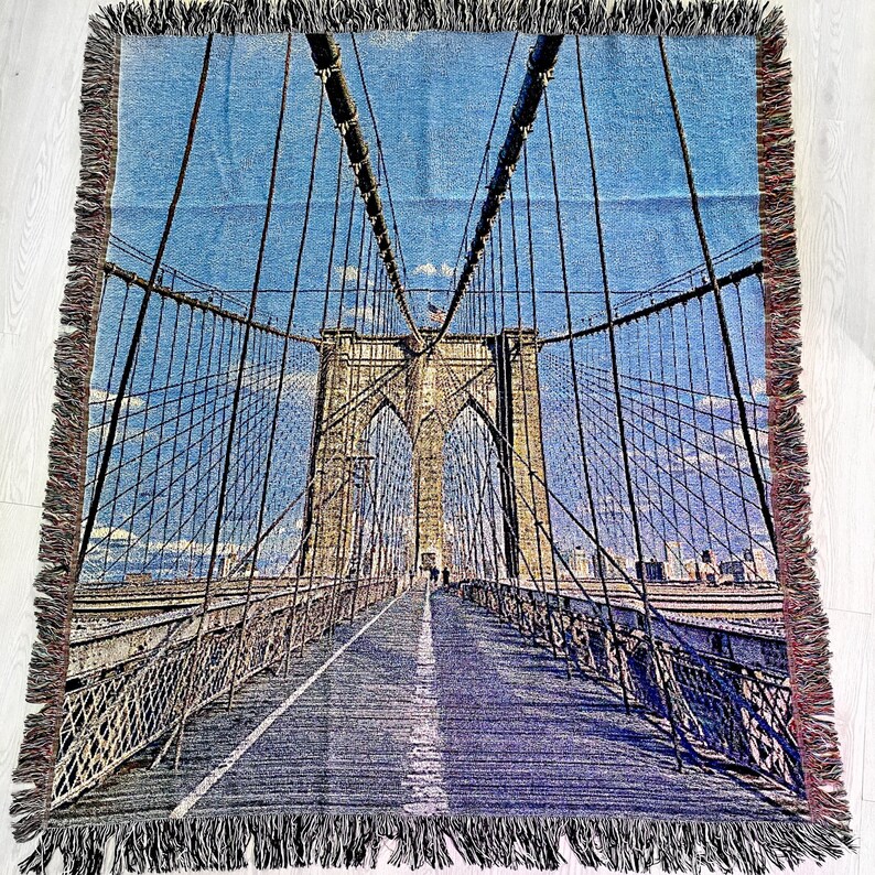 100% woven throw blanket with a photographic image of the Brooklyn Bridge in NYC. The image features a perspective view from the center of the footpath with an American flag on top of the bridge. The blanket has a colorful fringe on all sides.