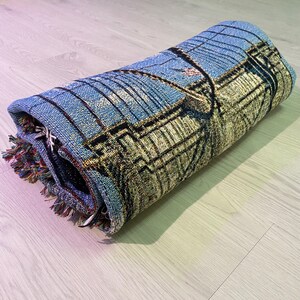 100% woven throw blanket with a photographic image of the Brooklyn Bridge in NYC. The image features a perspective view from the center of the footpath with an American flag on top of the bridge. The blanket has a colorful fringe on all sides.