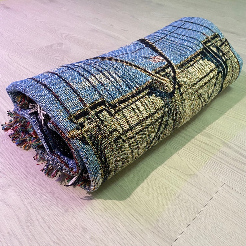 100% woven throw blanket with a photographic image of the Brooklyn Bridge in NYC. The image features a perspective view from the center of the footpath with an American flag on top of the bridge. The blanket has a colorful fringe on all sides.