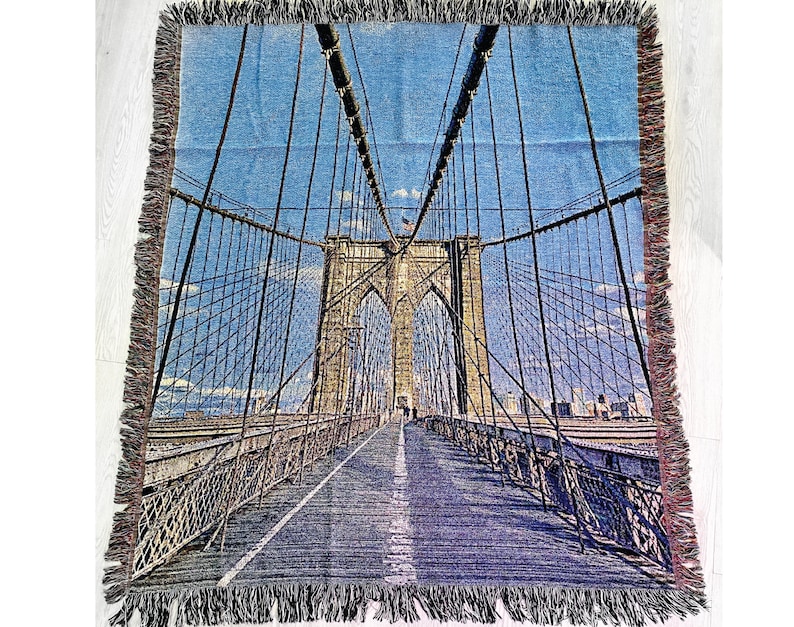 100% woven throw blanket with a photographic image of the Brooklyn Bridge in NYC. The image features a perspective view from the center of the footpath with an American flag on top of the bridge. The blanket has a colorful fringe on all sides.