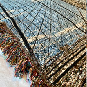 100% woven throw blanket with a photographic image of the Brooklyn Bridge in NYC. The image features a perspective view from the center of the footpath with an American flag on top of the bridge. The blanket has a colorful fringe on all sides.