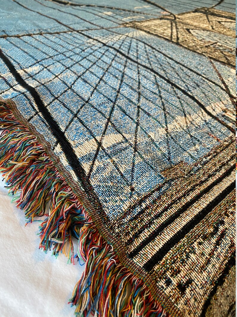 100% woven throw blanket with a photographic image of the Brooklyn Bridge in NYC. The image features a perspective view from the center of the footpath with an American flag on top of the bridge. The blanket has a colorful fringe on all sides.