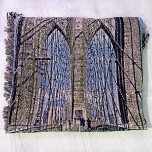 100% woven throw blanket with a photographic image of the Brooklyn Bridge in NYC. The image features a perspective view from the center of the footpath with an American flag on top of the bridge. The blanket has a colorful fringe on all sides.