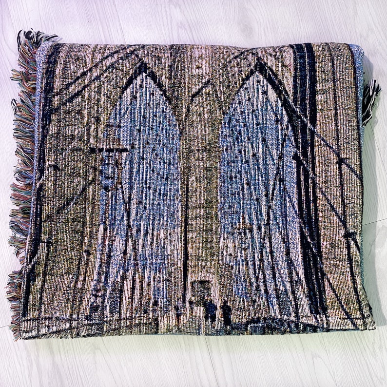 100% woven throw blanket with a photographic image of the Brooklyn Bridge in NYC. The image features a perspective view from the center of the footpath with an American flag on top of the bridge. The blanket has a colorful fringe on all sides.