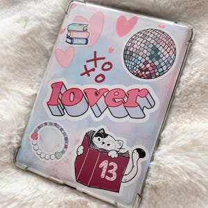 May include: Clear tablet case with various stickers. The stickers include a disco ball, hearts, books, a cat in a book with the number 13, and the word "lover". The case has a pink and blue watercolor background.