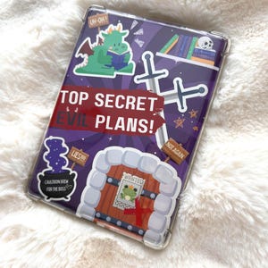 May include: A clear phone case with a purple design and cartoon graphics. The case features a green dragon reading a book, crossed swords, and the text "TOP SECRET EVIL PLANS!" Other graphics include a cauldron, a frog wanted poster, and the text "LIES!!!" and "NOT AGAIN."