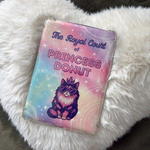 May include: A clear tablet case with a vibrant design. The case displays the text "The Royal Court of PRINCESS DONUT" and a cartoon cat wearing sunglasses and a crown. The background is a blend of pink, blue, and purple with star accents.