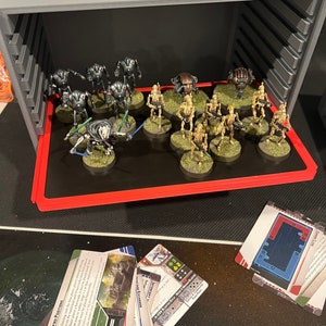 May include: A gray plastic storage case with a red tray inside. The tray holds a collection of miniature figures, including a group of black and gray figures and a group of tan figures. The figures are arranged in a grid pattern on the tray. There are also several cards on the table next to the case.