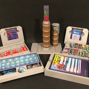 May include: A grey plastic organiser with multiple compartments for storing dice, cards, and tokens. The organiser has a tower in the centre with a stack of coloured blocks.