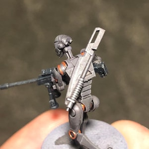 May include: A gray and orange robot miniature figure holding a gun and a silver rifle on its back. The figure is standing on a gray base.