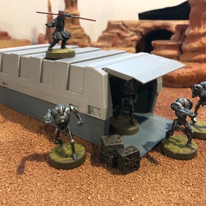 May include: A gray, rectangular, sci-fi themed building with an open door.  Four silver robots stand outside the building, and a red-clad figure stands on top of the building.