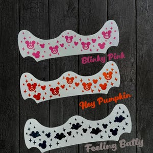 May include: Three white vinyl decals with different designs. The top decal features pink Mickey Mouse heads and stars. The middle decal features orange Mickey Mouse heads and stars. The bottom decal features black bats and Mickey Mouse heads with stars. The text on the decals reads "Blinky Pink", "Hey Pumpkin", and "Feeling Batty".