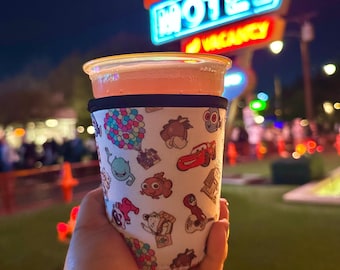 May include: A clear plastic cup filled with a pink beverage, held in front of a neon sign. The cup is wrapped in a white koozie printed with cartoon characters. The sign reads "Cozy Cone Motel" with a "Vacancy" sign.