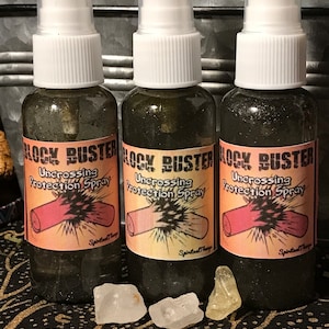 May include: Three glass bottles with black labels that read "Block Buster Uncrossing Protection Spray". The bottles are filled with a dark liquid and have white spray tops. The bottles are sitting on a black and gold patterned surface with three clear crystals.