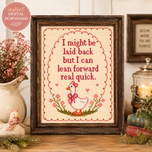 May include: A framed cross-stitch pattern on cream fabric featuring a white goose with a pink bow and the text: I might be laid back but I can lean forward real quick. The design is enclosed in a pink floral oval border with small hearts.