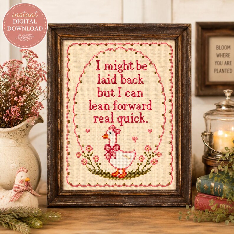 May include: A framed cross-stitch pattern on cream fabric featuring a white goose with a pink bow and the text: I might be laid back but I can lean forward real quick. The design is enclosed in a pink floral oval border with small hearts.