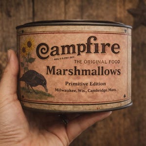 May include: A vintage-style tin of "Campfire Marshmallows" held in a hand. The label features a sunflower, a crow illustration, and text including "Primitive Edition" and city names. The tin has a rustic, aged appearance.