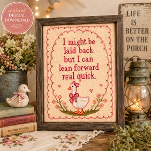 May include: A framed cross-stitch embroidery featuring a white goose with a pink bow, surrounded by a pink floral border and the text: I might be laid back but I can lean forward real quick. The piece is set in a dark brown wooden frame.
