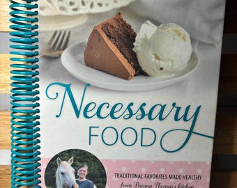 May include: A cookbook titled "Necessary Food: Traditional Favorites Made Healthy from Briana Thomas's Kitchen" with a teal and white cover. The cover features a slice of chocolate cake with vanilla ice cream, a white horse, and a woman.