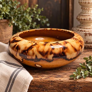 May include: A round ceramic bowl featuring a glossy finish with a marbled pattern of warm tan, golden yellow, and deep dark brown streaks. The bowl has a wide, curved rim and a smooth, reflective surface that catches light across its rounded exterior.