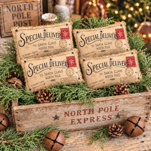 May include: Four rectangular beige fabric pillows featuring a vintage-style Special Delivery print addressed to Santa Claus at the North Pole. The pillows are nestled in a weathered wooden crate labeled North Pole Express with metal jingle bells.