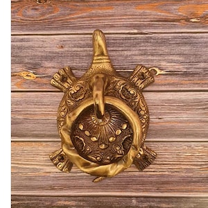 May include: A brass turtle-shaped door knocker. The knocker features a ring for knocking and is decorated with intricate patterns. The turtle's head and flippers are detailed, and the overall design has an antique appearance.