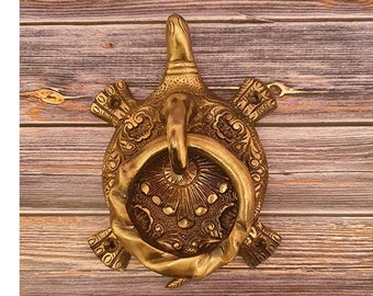 May include: A brass turtle-shaped door knocker. The knocker features a ring for knocking and is decorated with intricate patterns. The turtle's head and flippers are detailed, and the overall design has an antique appearance.