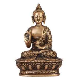 Puede incluir: Una estatua de Buda de color bronce en posición sentada, con una mano levantada en un gesto de bendición. La estatua presenta intrincados detalles en las túnicas y la base, sobre un fondo blanco. La estatua es una representación del arte budista.