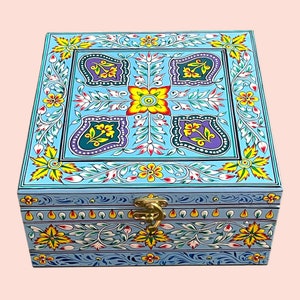 May include: A square, hand-painted wooden box with a light blue background and floral designs in yellow, purple, and green. The box has a brass clasp and intricate patterns, showcasing a detailed, artistic aesthetic. The box is a decorative storage item.