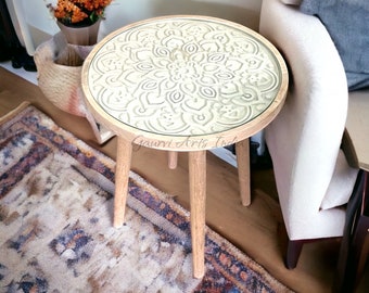 Puede incluir: Una mesa auxiliar redonda con una tapa de cristal con un diseño floral tallado. La mesa tiene un marco de madera clara y tres patas. La mesa mide aproximadamente 51 cm de diámetro y 61 cm de altura.