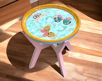 Puede incluir: Una mesa auxiliar redonda con una tapa azul claro con diseños de mariposas y flores. La mesa tiene un borde amarillo y dorado, y patas rosas. La mesa tiene aproximadamente 46 cm de diámetro y 46 cm de alto.