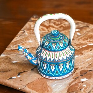 May include: A hand-painted, decorative teapot with a white handle and a blue body. The teapot features intricate floral patterns in white, green, and orange. The spout and lid are also adorned with similar designs. The teapot sits on a brown surface.