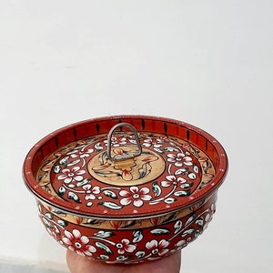 May include: A hand-painted, round, lidded container with a red background and white floral design. The lid has a metal handle. The container is held in a hand against a white background.