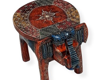 May include: A handcrafted wooden elephant stool with a round top. The stool is painted in shades of red, blue, and white, with intricate patterns on the top and legs. The elephant's head forms the front of the stool, adding a unique design element.