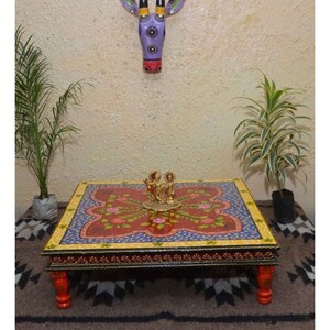 May include: A hand-painted wooden coffee table with a floral design in red, blue, and yellow. The table has red legs and a decorative border. A small brass statue sits on top. The table is a unique piece of home decor.